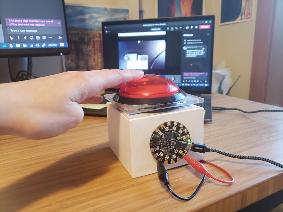 A white box with a big red button on top with a hand about to press the button. On the front of the bix is a circuit playground express with crocodile clips connecting it to the white box. In the background is a laptop showing a Microsoft teams meeting